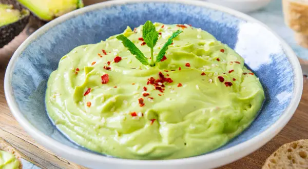 Cremige Avocado-Creme, garniert mit Minze, Sesam und Chiliflocken, serviert in einer blauen Schüssel.