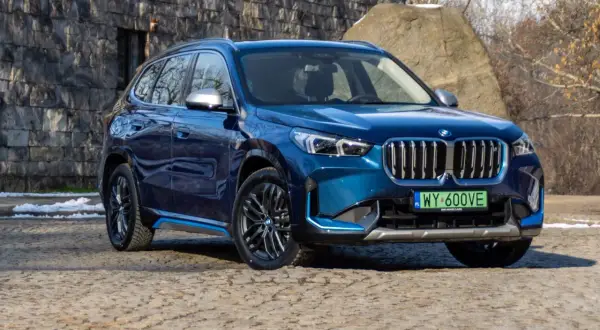 Błękitny BMW X1 elektryk z niebieskimi akcentami na progu, zaparkowany na kamienistej nawierzchni.