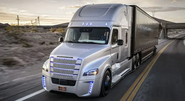 Un camion américain nez plat argenté roule sur une route déserte au coucher du soleil.