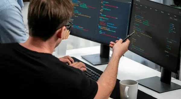 Dwóch programistów analizuje kod na monitorach. To świetny przykład, jak wygląda praca w IT dla początkujących – nauka i współpraca.