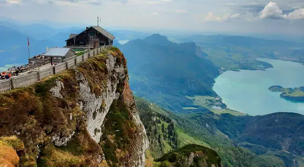 Ein Gasthaus thront am Felsvorsprung, die Himmelspforte am Schafberg, mit Blick auf einen türkisfarbenen See und grüne Berge.
