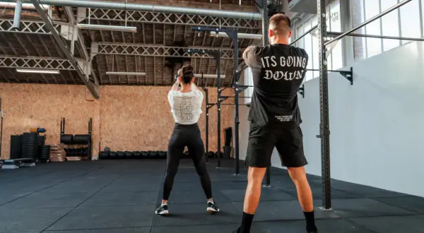 Para osiągnięcia dobrych wyników w crossfit, jak często trenować? Para ćwiczy w siłowni, kobieta z kettlebell, mężczyzna w koszulce "It's Going Down".