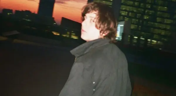 Francesco De Leo, con i capelli castani mossi, indossa un cappotto scuro. Sullo sfondo, un tramonto arancione illumina i profili degli edifici urbani.