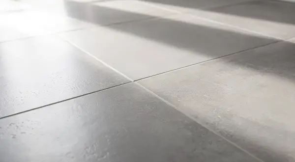 Lumière du soleil traversant un carrelage gris, créant des ombres douces. On entendrait presque le carrelage qui sonne creux sous les pas.