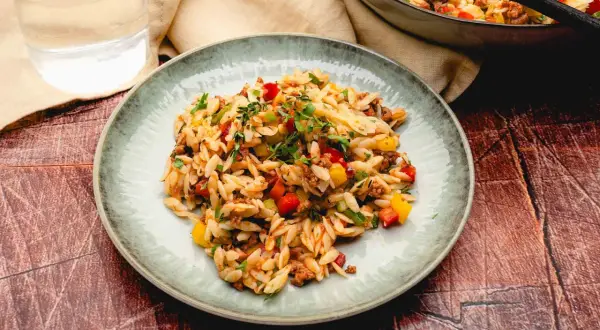 Ein Teller mit kritharaki salat mit hackfleisch, garniert mit frischen Kräutern. Bunte Paprikawürfel und Hackfleisch sind unter die Nudeln gemischt.