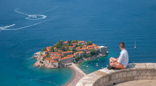 Czarnogóra na co uważać? Widok na wyspę Sveti Stefan, turysta kontempluje morze. Uważaj na słońce i tłumy na plaży.