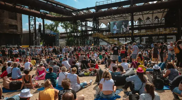 Tłum ludzi na festiwalu muzycznym, siedzących na ziemi i słuchających koncertu. W tle widać scenę i wielki ekran z występującymi artystami. To część owf lineup.