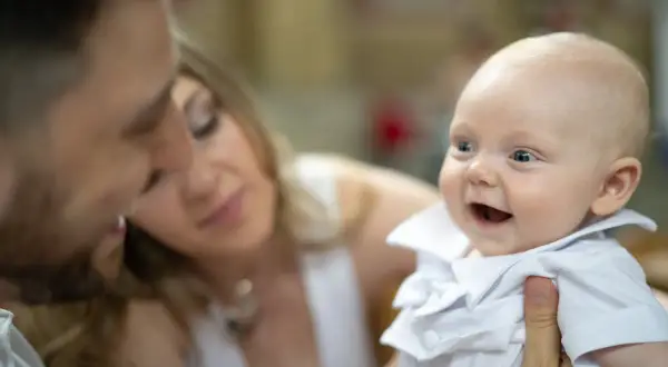 Un bébé souriant en tenue blanche, entouré de ses parents. Un moment précieux pour un baptême civil, depuis quand cette tradition existe-t-elle ?