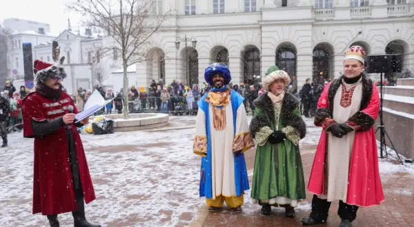 Król Melchior w niebieskim stroju z turbanem, obok innych postaci w barwnych szatach, podczas zimowego wydarzenia.
