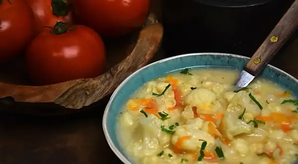 Pyszna zupa kalafiorowa z makaronem, ozdobiona natką pietruszki. Obok miski leżą świeże pomidory.
