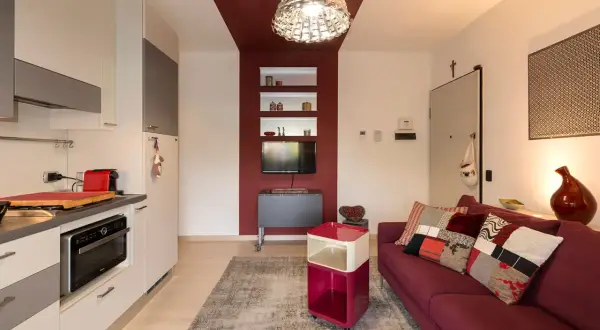 Soggiorno con divano bordeaux, tappeto grigio e una parete bordeaux e tortora che incornicia una nicchia con TV e mensole.