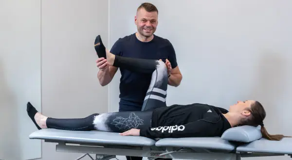 Fizjoterapeuta pomaga pacjentce w ćwiczeniach rehabilitacja kolana po urazie. Kobieta leży na kozetce, mężczyzna delikatnie zgina jej nogę.