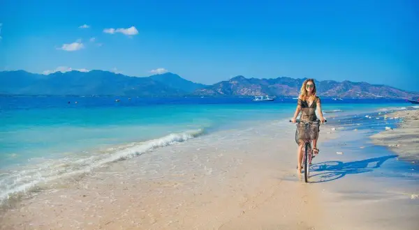 Kobieta na rowerze przemierza piaszczystą plażę z widokiem na turkusowe morze i góry. To idylliczny krajobraz, jak z marzeń o podróży na półwysep Indochiński.