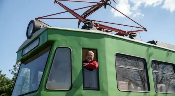 Uśmiechnięty mężczyzna w czerwonym swetrze wygląda z okna zielonej lokomotywy. To historyczna łódź Skierniewice, gotowa do podróży.