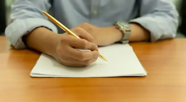 Main tenant une plume dorée, une personne rédige une lettre de motivation STAPS sur une feuille blanche.