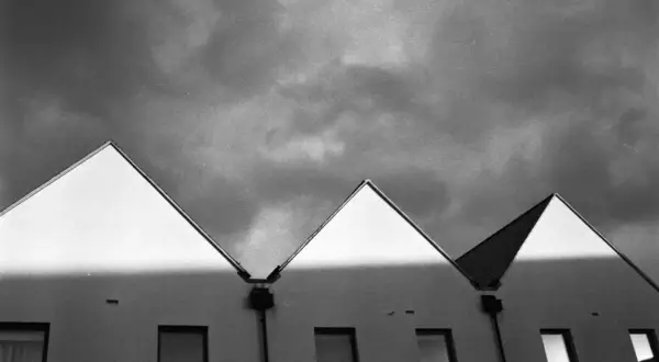 Row of modern houses with sharp, triangular roofs against a dramatic, cloudy sky. The light creates a stark contrast, highlighting the architectural lines, a testament to careful stand development.