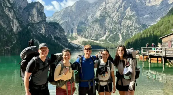 Fem glada vandrare med stora ryggsäckar njuter av en vacker sjö och berg i Italien.