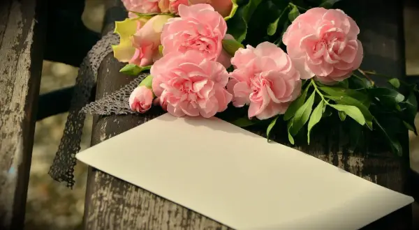 Bukiet różowych kwiatów, koronka i pusta kartka na drewnianym tle.