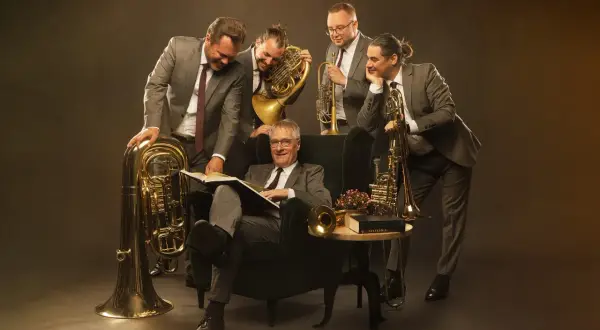 Fünf Musiker mit Blasinstrumenten, darunter Tuba, Horn und Trompete, umringen einen Mann im Sessel, der ein Buch liest.