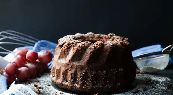 En saftig sockerkaka choklad, pudrad med florsocker, med vindruvor och visp i bakgrunden.