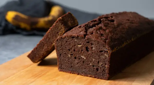 En saftig banankaka med choklad, en skiva är uppskuren. I bakgrunden syns en övermogen banan.