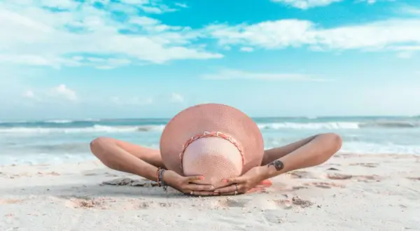 Osoba w kapeluszu leży na plaży, ręce za głową. To idealny przykład, co to znaczy last minute – spontaniczny relaks nad morzem.