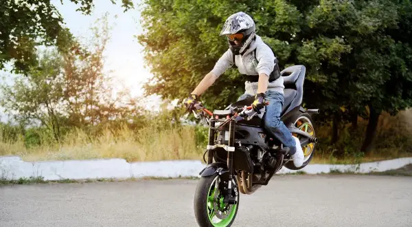 Motocyklista w kasku wykonuje jazdę na tylnym kole na asfalcie, otoczony zielenią.