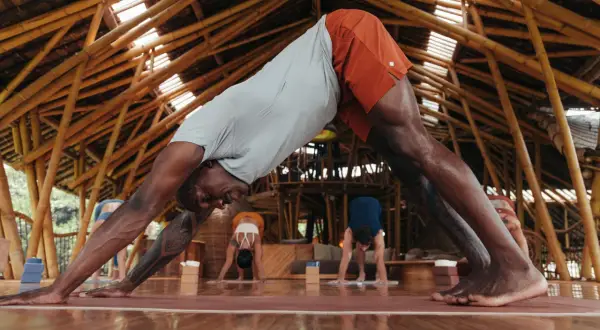 Un homme en posture de chien tête en bas, cherchant à redresser son dos. D'autres pratiquent le yoga dans un studio en bambou.