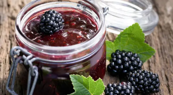 Glas mit Brombeermarmelade, daneben frische Brombeeren und Blätter. Perfekt, um sie mit Joghurt oder auf Toast zu mischen.