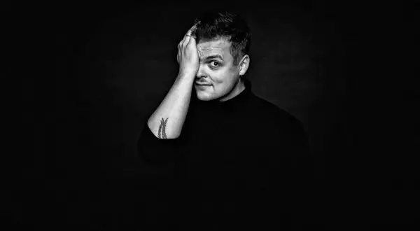 Czarno-białe zdjęcie. Mężczyzna z tatuażem na ramieniu, w czarnym golfie, zasłaniający dłonią jedno oko. Wygląda jak król piosenkarz.