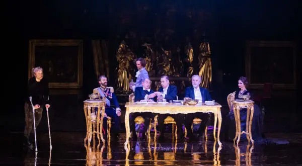 Scena z "Dziadów" w warszawskim teatrze. Przy złotym stole siedzą mężczyźni w strojach z epoki, obok kobieta na wózku.