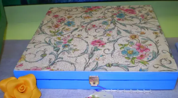 Caja azul con tapa decorada con papel decoupage floral. Un detalle de rosa amarilla y etiqueta floral completan la escena.