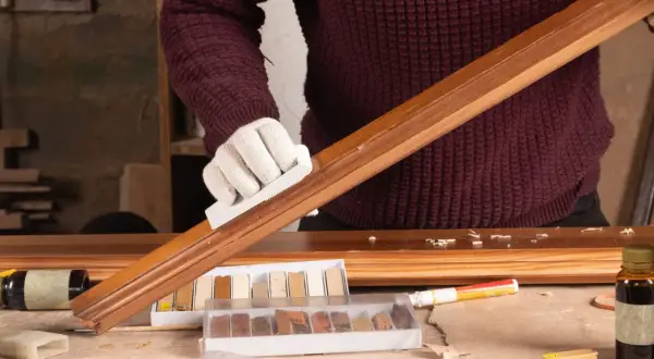 Persona con guantes blancos restaura un mueble de madera, usando un bloque para lijar y kits de retoque.