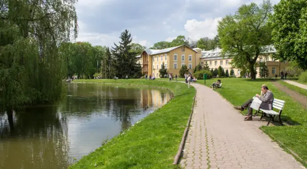 Piękny park przy sanatorium z ZUS. Spacerowicze odpoczywają nad stawem, a w oddali widać żółty budynek.