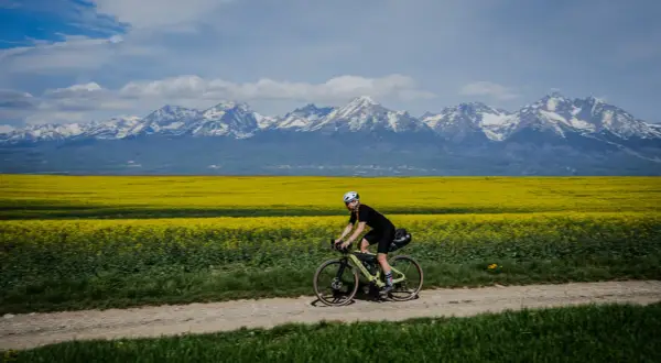 Rowerzystka przemierza polną drogę na tle kwitnącego rzepaku i ośnieżonych Tatr. Marzenie o rowerem dookoła Tatr w jeden dzień staje się rzeczywistością.