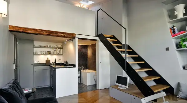 Interno moderno con cucina a vista, bagno piastrellato e scala a giorno. Le case con soffitti alti sfruttano lo spazio verticale con un soppalco visibile.