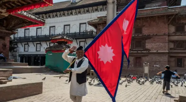 Mężczyzna macha flagą Nepalu na placu pełnym gołębi. W tle tradycyjna architektura.