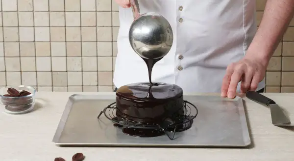 Ein Konditor überzieht einen Kuchen mit glänzender mirror glaze. Schokoladenstückchen liegen daneben.