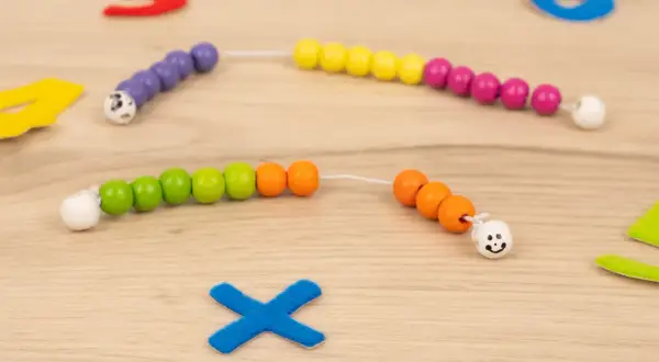 Bunte Perlenketten zum **rechenkette basteln** liegen auf Holz. Zahlen und ein blaues Kreuz sind im Hintergrund.
