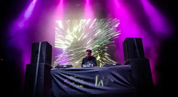 DJ na scenie, fioletowe światła, wizualizacje na ekranie. Sprawdź audioriver timetable, by nie przegapić jego występu!