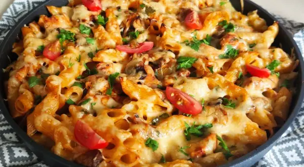 Pyszna zapiekanka makaronowa z kurczakiem, okraszona roztopionym serem, pomidorkami i natką pietruszki, podana w okrągłej patelni.