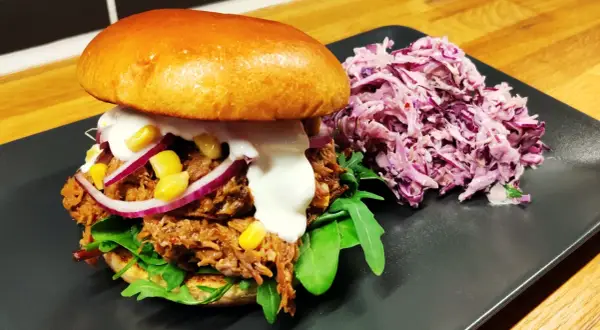 En saftig pulled pork-burgare med majs, rödlök och krämig sås, serverad med en lila kålsallad.