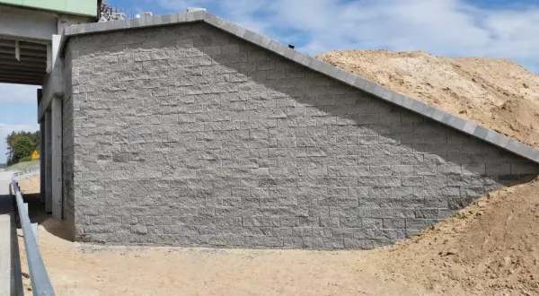 Mur oporowy z szarej kostki brukowej, obok nasypu ziemnego i fragmentu wiaduktu.
