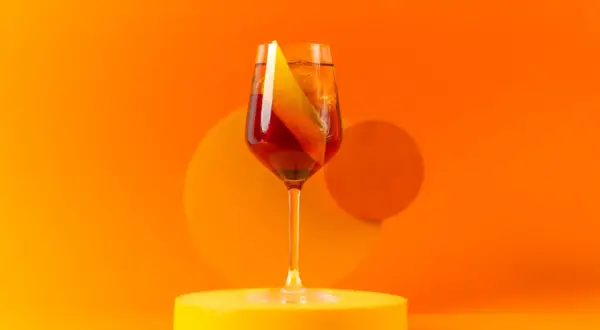 Ein Glas Aperitif mit Orangenscheibe auf gelber Unterlage vor orangefarbenem Hintergrund.