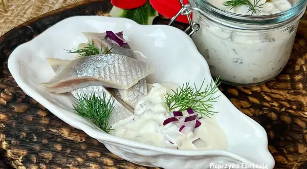 Delikatne śledzie w śmietanie z koperkiem i czerwoną cebulką, podane w białej miseczce. Obok słoiczek z sosem.