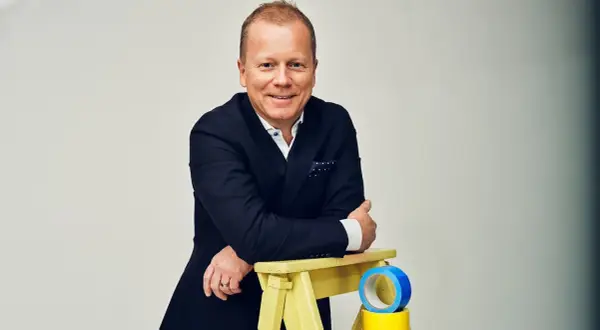 Claes Seldeby, en glad man i kostym, lutar sig mot en gul pall med blå och gul tejp.