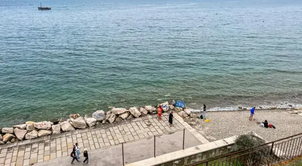 Spacerem po wybrzeżu w Słowenii Piran. Widać łódź na morzu i kilka osób odpoczywających na kamienistej plaży.