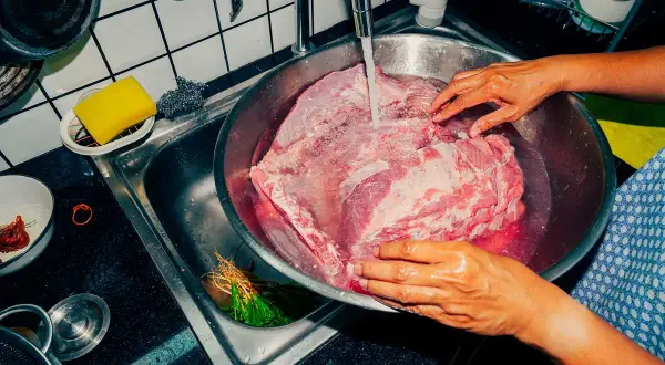 Ręce płuczą duży kawałek wieprzowiny pod bieżącą wodą. To ważny krok, by dowiedzieć się, ile gotować wieprzowinę.