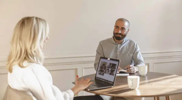 Två personer vid ett runt bord, en kvinna med blont hår och en man med skägg. De har en laptop och anteckningsblock, vilket tyder på ett konstruktivt samtal.