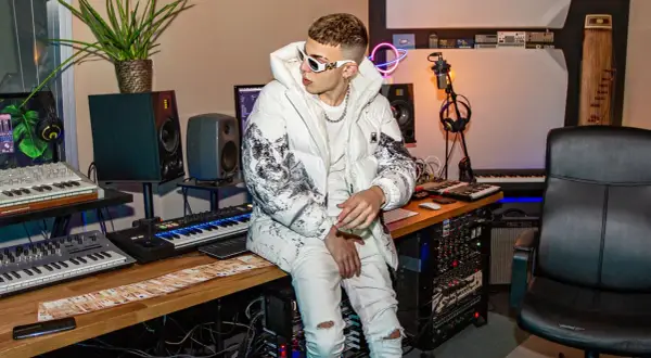 Un giovane skinny cantante con occhiali bianchi e giacca imbottita è seduto in uno studio di registrazione, circondato da tastiere e apparecchiature audio.